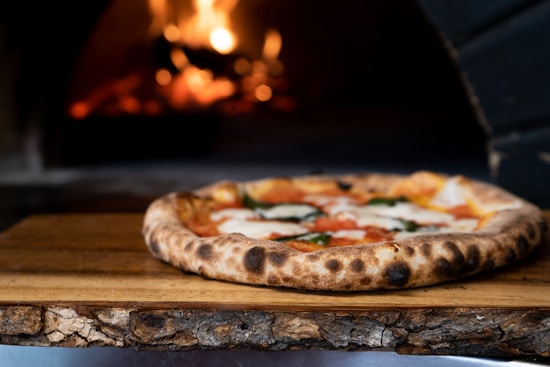 A wood-fired pizza with a charred crust sits on a rustic wooden board. The pizza is topped with melted cheese, tomato sauce, and fresh basil. In the background, a fire is burning in a stone oven, casting a warm glow.