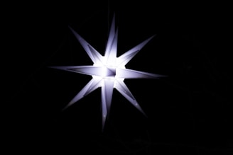 A bright, multi-pointed star-shaped light fixture glows against a pitch-black background. The star has a symmetrical design with sharp, elongated points extending outward in all directions.