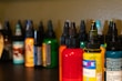 Several bottles of colored ink are arranged on a dark surface, each with a pointed cap. The inks are in various colors including red, yellow, orange, green, and more with labels on each bottle. The background is a neutral shade, slightly out of focus, allowing the vibrant colors of the bottles to stand out.