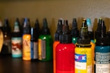 Several bottles of colored ink are arranged on a dark surface, each with a pointed cap. The inks are in various colors including red, yellow, orange, green, and more with labels on each bottle. The background is a neutral shade, slightly out of focus, allowing the vibrant colors of the bottles to stand out.