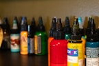 Several bottles of colored ink are arranged on a dark surface, each with a pointed cap. The inks are in various colors including red, yellow, orange, green, and more with labels on each bottle. The background is a neutral shade, slightly out of focus, allowing the vibrant colors of the bottles to stand out.