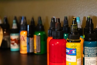 Several bottles of colored ink are arranged on a dark surface, each with a pointed cap. The inks are in various colors including red, yellow, orange, green, and more with labels on each bottle. The background is a neutral shade, slightly out of focus, allowing the vibrant colors of the bottles to stand out.