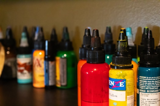Several bottles of colored ink are arranged on a dark surface, each with a pointed cap. The inks are in various colors including red, yellow, orange, green, and more with labels on each bottle. The background is a neutral shade, slightly out of focus, allowing the vibrant colors of the bottles to stand out.