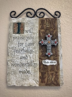 A decorative wall plaque featuring a religious inscription on a stone-like surface. The text reads: 'I will praise you for I am fearfully and wonderfully made.' To the right, there is an ornamental metal cross with red accents on a patterned fabric background. The reference 'Psalm 139:14' is inscribed beneath the cross. The plaque is hung on a wall using a black ornamental bracket.