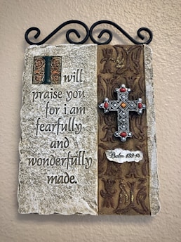 A decorative wall plaque featuring a religious inscription on a stone-like surface. The text reads: 'I will praise you for I am fearfully and wonderfully made.' To the right, there is an ornamental metal cross with red accents on a patterned fabric background. The reference 'Psalm 139:14' is inscribed beneath the cross. The plaque is hung on a wall using a black ornamental bracket.