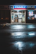 The front entrance of a family drug store named 'Alpha Pharmacy' at night, illuminated by bright, artificial lighting. The pavement in front of the store is wet, reflecting the store's lights. There is a sign above the entrance displaying 'Family Drug' prominently.