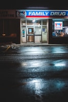 The front entrance of a family drug store named 'Alpha Pharmacy' at night, illuminated by bright, artificial lighting. The pavement in front of the store is wet, reflecting the store's lights. There is a sign above the entrance displaying 'Family Drug' prominently.