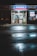 The front entrance of a family drug store named 'Alpha Pharmacy' at night, illuminated by bright, artificial lighting. The pavement in front of the store is wet, reflecting the store's lights. There is a sign above the entrance displaying 'Family Drug' prominently.