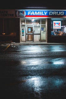 The front entrance of a family drug store named 'Alpha Pharmacy' at night, illuminated by bright, artificial lighting. The pavement in front of the store is wet, reflecting the store's lights. There is a sign above the entrance displaying 'Family Drug' prominently.