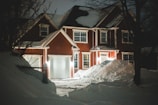 A two-story brick house with a steep, snow-covered roof. The house features large white-framed windows and a white garage door. The front yard is covered with deep snowdrifts, illuminated by warm porch lights at night. Bare trees and a fence are also visible around the house.