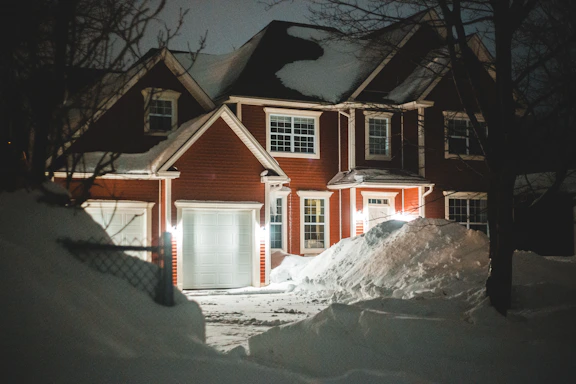 A two-story brick house with a steep, snow-covered roof. The house features large white-framed windows and a white garage door. The front yard is covered with deep snowdrifts, illuminated by warm porch lights at night. Bare trees and a fence are also visible around the house.