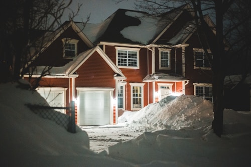 A two-story brick house with a steep, snow-covered roof. The house features large white-framed windows and a white garage door. The front yard is covered with deep snowdrifts, illuminated by warm porch lights at night. Bare trees and a fence are also visible around the house.