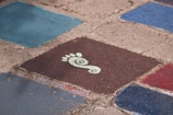 Several colorful, square tiles with rough, textured surfaces arranged on the ground. One tile prominently features a stylized white footprint with a spiral design on a dark brown background. The surrounding tiles are shades of blue and red.