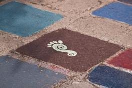 Several colorful, square tiles with rough, textured surfaces arranged on the ground. One tile prominently features a stylized white footprint with a spiral design on a dark brown background. The surrounding tiles are shades of blue and red.