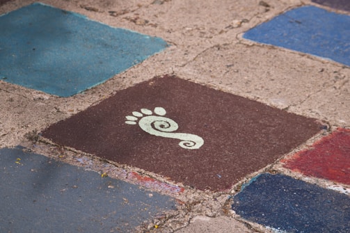 Several colorful, square tiles with rough, textured surfaces arranged on the ground. One tile prominently features a stylized white footprint with a spiral design on a dark brown background. The surrounding tiles are shades of blue and red.