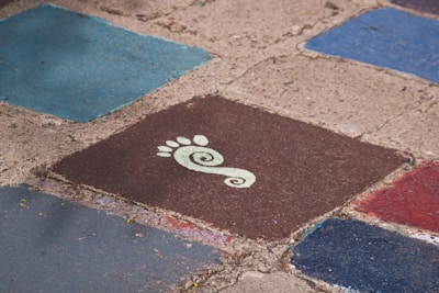 Several colorful, square tiles with rough, textured surfaces arranged on the ground. One tile prominently features a stylized white footprint with a spiral design on a dark brown background. The surrounding tiles are shades of blue and red.