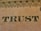 The word 'TRUST' is prominently displayed on a textured beige surface, featuring intricate, dark, serif typography. Above the word, there is a decorative pattern consisting of interlocking loops and circles, resembling ornamental engraving typically found on paper currency.