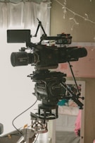 A professional film camera is mounted on a complex rig setup in an indoor environment. The camera has multiple components, including lenses and adjustment knobs, and is aligned with a visible tripod or stabilizer. The background shows a partially visible room with a curtained window, some wall decorations, and a hint of furnishings.