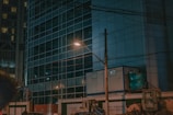 A nighttime urban scene with a modern glass-fronted building in the background. There is a streetlamp illuminating part of the scene and casting a warm light. In the foreground, construction equipment and temporary structures can be seen. Electrical wires are visible, crossing the street above.