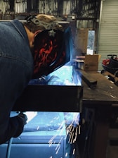 A person is engaged in welding, wearing a protective helmet with flame designs. Bright sparks are flying around, and a blue light emits from the welding area. The setting appears to be an industrial workshop with metallic surfaces and various equipment in the background.