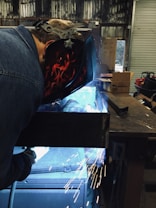 A person is engaged in welding, wearing a protective helmet with flame designs. Bright sparks are flying around, and a blue light emits from the welding area. The setting appears to be an industrial workshop with metallic surfaces and various equipment in the background.