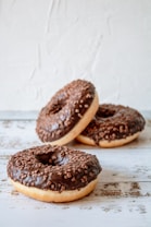 Three chocolate-glazed donuts with sprinkles are arranged on a rustic wooden surface. The front donut is prominently displayed, with the others slightly blurred in the background. The texture of the sprinkles and the smoothness of the glaze are visibly appealing.