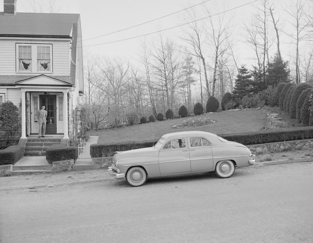 grayscale photo of vintage car parked beside house