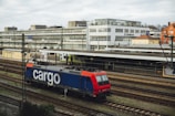A freight train with a blue and red locomotive marked with the word 'cargo' is situated on railway tracks in an urban setting. The scene includes a large building in the background with multiple windows, and an overcast sky adds to the atmosphere.
