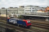 A freight train with a blue and red locomotive marked with the word 'cargo' is situated on railway tracks in an urban setting. The scene includes a large building in the background with multiple windows, and an overcast sky adds to the atmosphere.