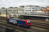 A freight train with a blue and red locomotive marked with the word 'cargo' is situated on railway tracks in an urban setting. The scene includes a large building in the background with multiple windows, and an overcast sky adds to the atmosphere.
