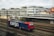 A freight train with a blue and red locomotive marked with the word 'cargo' is situated on railway tracks in an urban setting. The scene includes a large building in the background with multiple windows, and an overcast sky adds to the atmosphere.