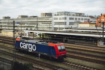 A freight train with a blue and red locomotive marked with the word 'cargo' is situated on railway tracks in an urban setting. The scene includes a large building in the background with multiple windows, and an overcast sky adds to the atmosphere.