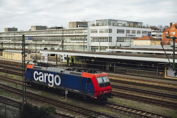 A freight train with a blue and red locomotive marked with the word 'cargo' is situated on railway tracks in an urban setting. The scene includes a large building in the background with multiple windows, and an overcast sky adds to the atmosphere.