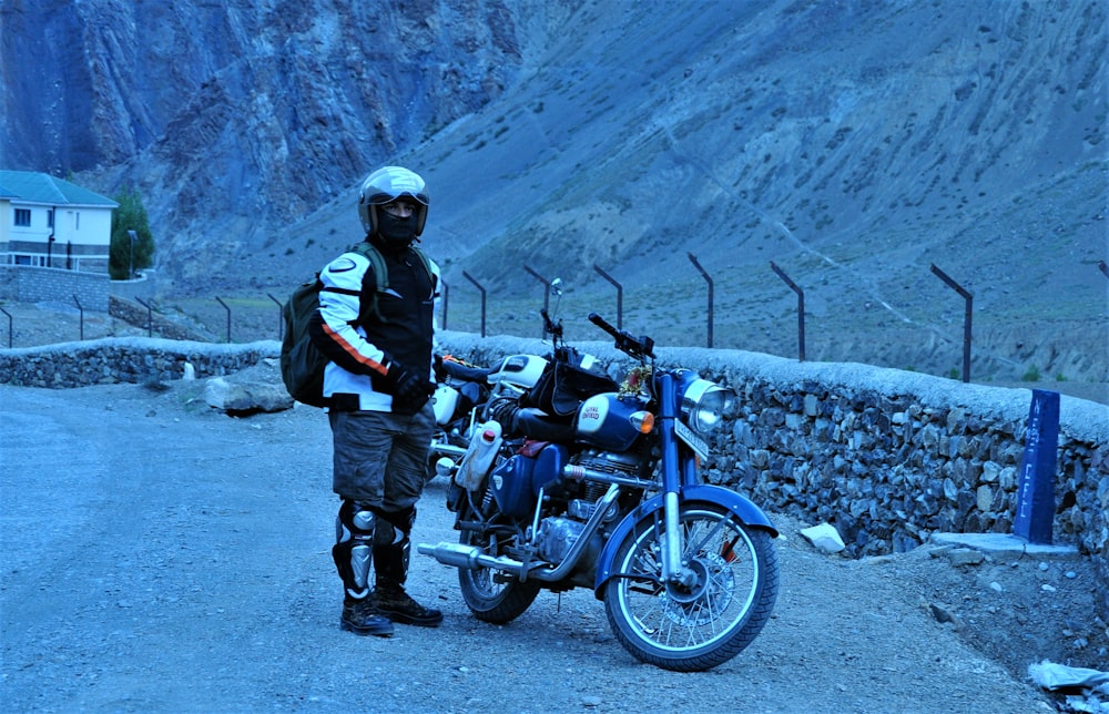 검은 재킷과 검은 바지를 입은 남자가 낮에 파란색 오토바이 옆에 서 있습니다.
