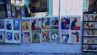 Several vintage posters are displayed on a street wall in an outdoor setting. The posters feature various designs, including advertisements for entertainment venues, food products, and art. The posters include vibrant artwork and illustrations, with text in French. The surrounding area also has some framed pictures and postcards displayed for sale.