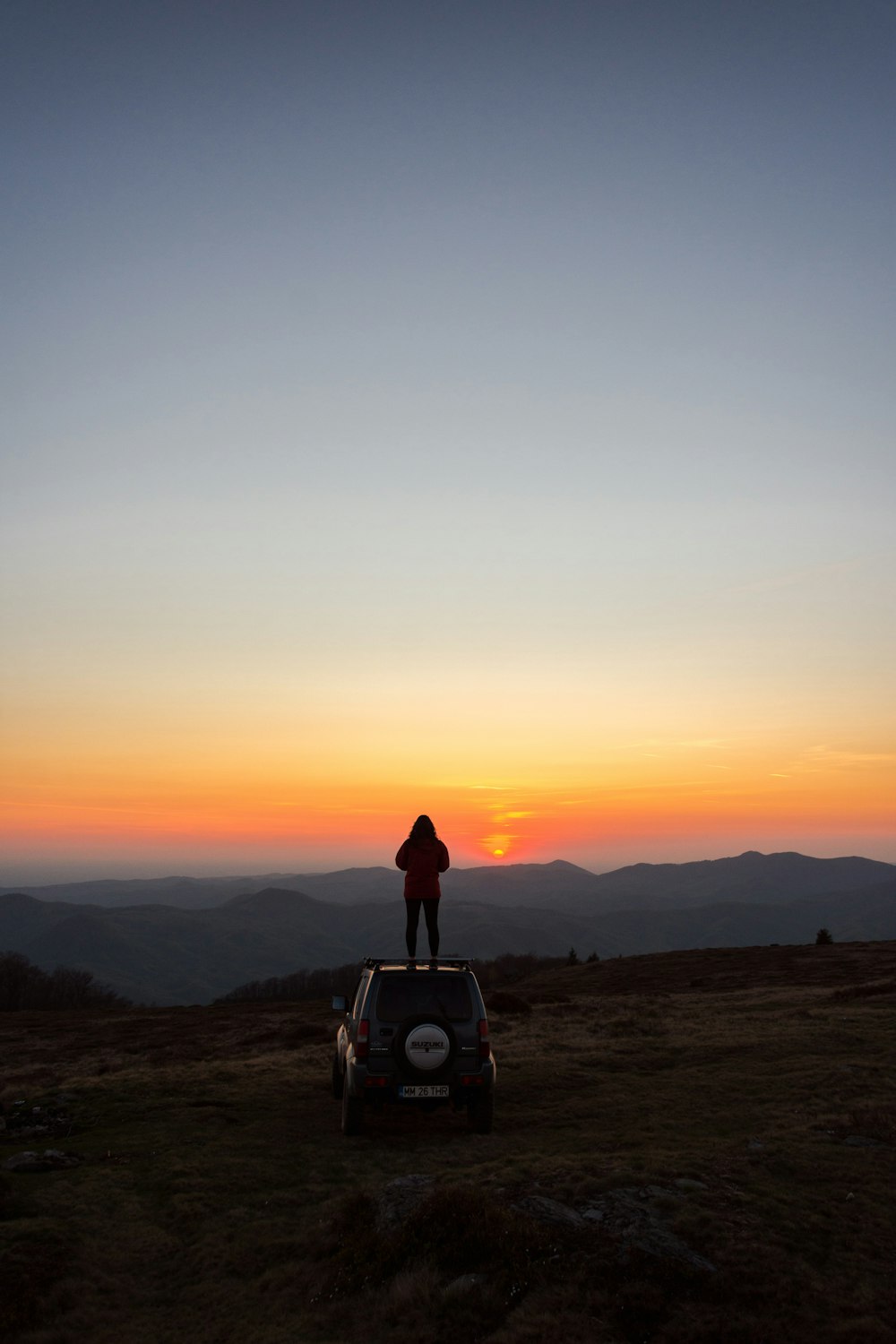 man in black jacket standing on top of the car during sunset