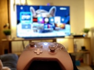 A gaming controller held in the foreground with a blurred television displaying a home screen interface in the background. The room has a cozy atmosphere with plants and a wooden shelf.