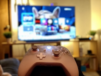 A gaming controller held in the foreground with a blurred television displaying a home screen interface in the background. The room has a cozy atmosphere with plants and a wooden shelf.