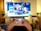 A gaming controller held in the foreground with a blurred television displaying a home screen interface in the background. The room has a cozy atmosphere with plants and a wooden shelf.