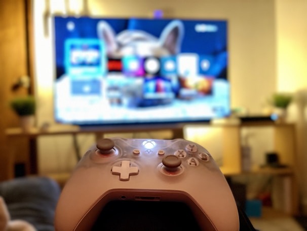 A gaming controller held in the foreground with a blurred television displaying a home screen interface in the background. The room has a cozy atmosphere with plants and a wooden shelf.
