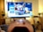 A gaming controller held in the foreground with a blurred television displaying a home screen interface in the background. The room has a cozy atmosphere with plants and a wooden shelf.