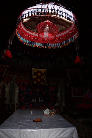 A traditional interior with a richly decorated ceiling featuring red and blue patterns and hanging tassels. Below, a table is set with a white cloth, a tray of oranges, a teapot, and a small bowl. The walls are adorned with vividly colored textiles and two animal pelts.