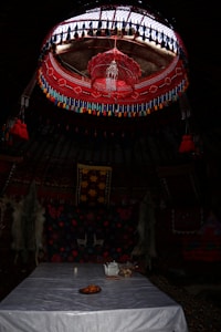 A traditional interior with a richly decorated ceiling featuring red and blue patterns and hanging tassels. Below, a table is set with a white cloth, a tray of oranges, a teapot, and a small bowl. The walls are adorned with vividly colored textiles and two animal pelts.