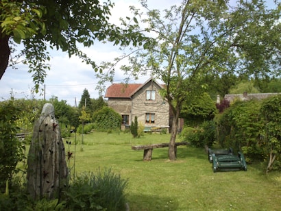 A rustic stone cottage with a red roof sits in a lush, green garden. The garden features various trees and shrubs, a wooden bench, and a stone sculpture adorned with floral patterns. A lawn mower is parked on the neatly manicured grass, and the sky is partly cloudy.
