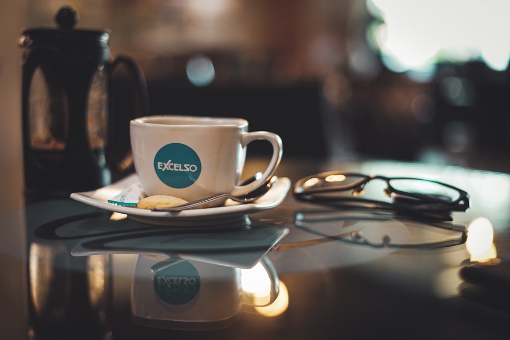 white ceramic mug on saucer beside black framed eyeglasses