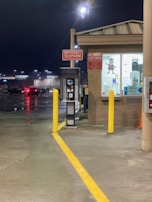 An emergency telephone is positioned next to a small building with illuminated signage. A 'No Smoking, Stop Engine' sign is also visible. The setting appears to be a parking area at night, as indicated by parked cars and streetlights reflecting off the wet pavement.