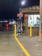 An emergency telephone is positioned next to a small building with illuminated signage. A 'No Smoking, Stop Engine' sign is also visible. The setting appears to be a parking area at night, as indicated by parked cars and streetlights reflecting off the wet pavement.