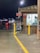 An emergency telephone is positioned next to a small building with illuminated signage. A 'No Smoking, Stop Engine' sign is also visible. The setting appears to be a parking area at night, as indicated by parked cars and streetlights reflecting off the wet pavement.