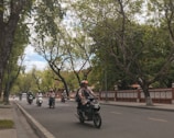 Motorcyclists ride along a tree-lined street. The road is bordered by a low red and white wall, with lush green trees and clear blue skies creating a serene backdrop.