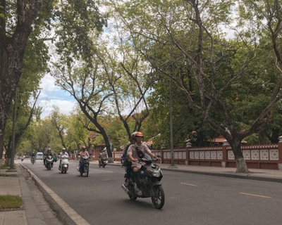 Motorcyclists ride along a tree-lined street. The road is bordered by a low red and white wall, with lush green trees and clear blue skies creating a serene backdrop.